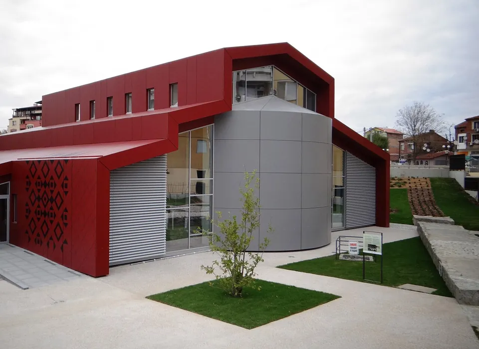 A modern building of red and grey panels with a large window and a cylindrical grey tower, set in a small landscaped park with lawns and young trees.