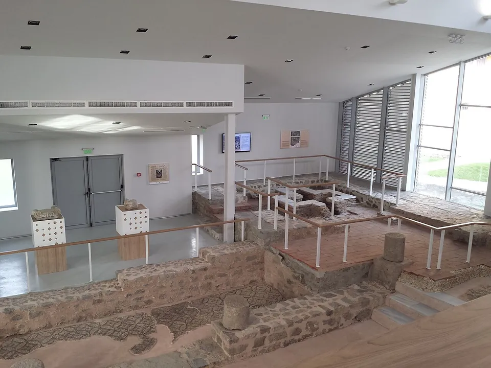 Inside a bright modern hall, the low stone walls of an early church and patches of geometric floor mosaic lie below railed walkways and glass, with display cases along the white walls.