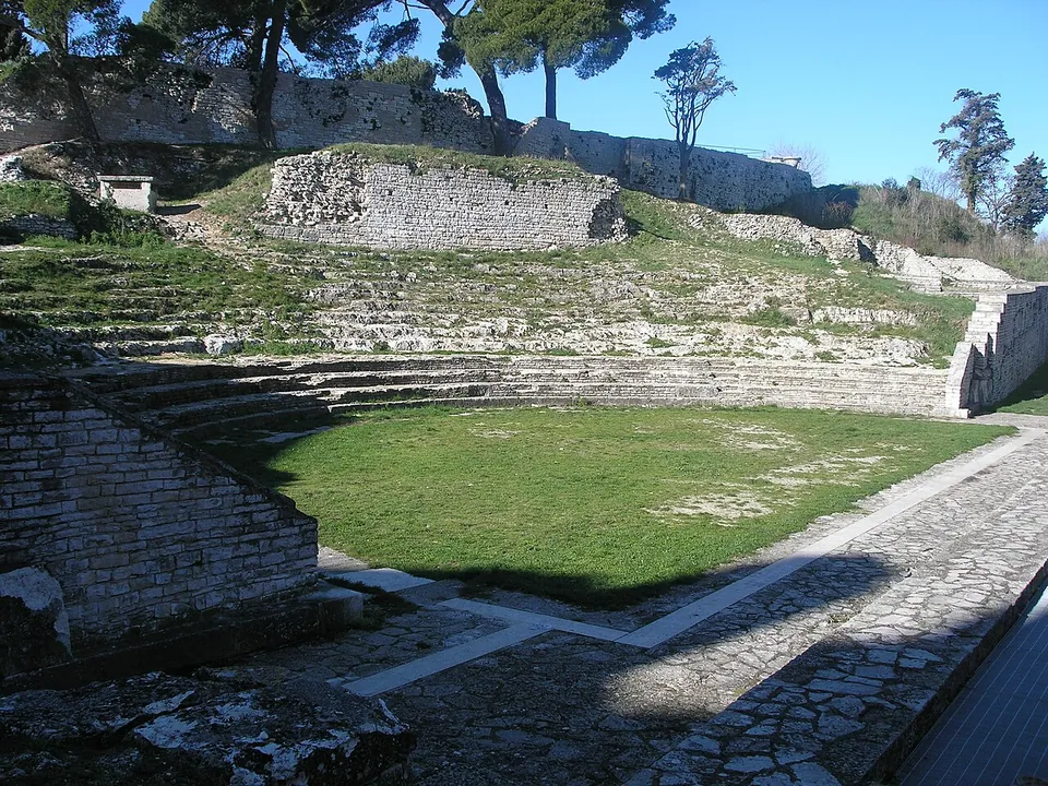 Tiers of stone seats rising in a curve up a hillside behind a green lawn, with the paved stage area and its low walls in the foreground and pine trees on the ridge above.