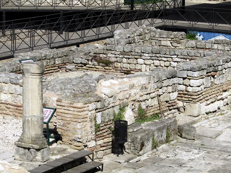 Low walls of stone with brick courses and a single re-erected stone column in an excavated area, a metal railing and walkway behind.