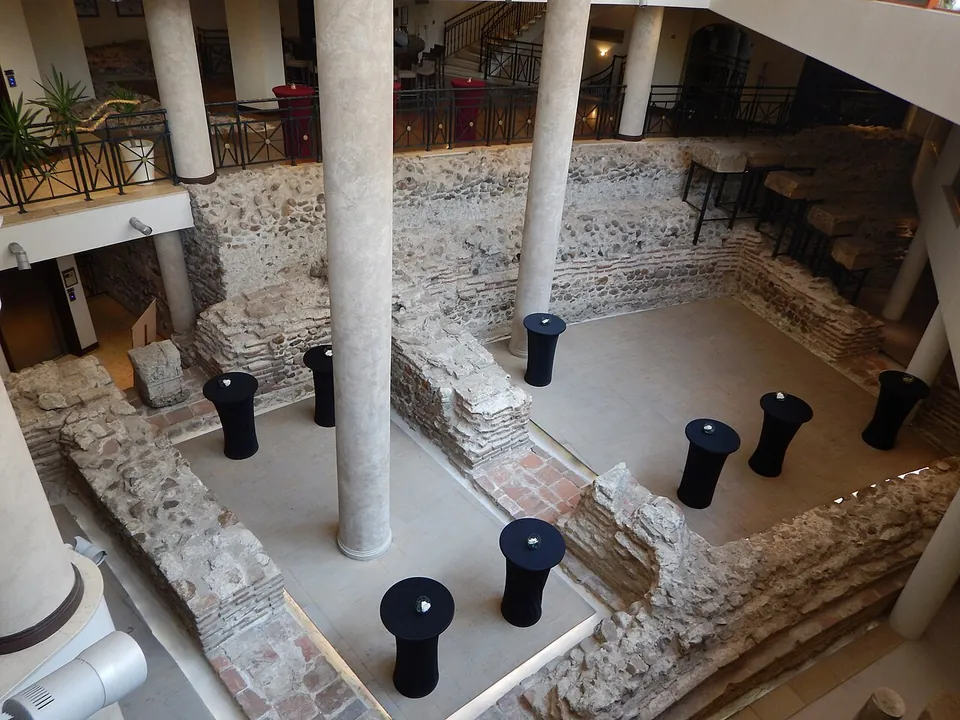 Rubble-stone walls with courses of brick, the lower part of the amphitheatre, laid out across the floor of a hotel atrium among tall modern columns, with black cocktail tables set between the walls and a balcony railing above.