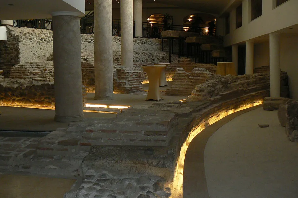 A curved wall of stone and brick lit from below by a strip of warm light, running round a pale modern floor, with a column and higher walls behind it in a dim interior.