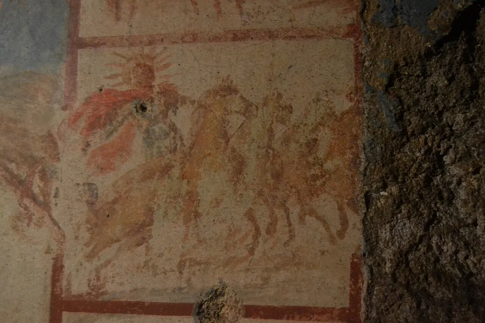 A close view of one small painted panel inside a red frame, faded to pale ochre and red, showing a radiate head and the outlines of horses beside the rough rock of the wall.