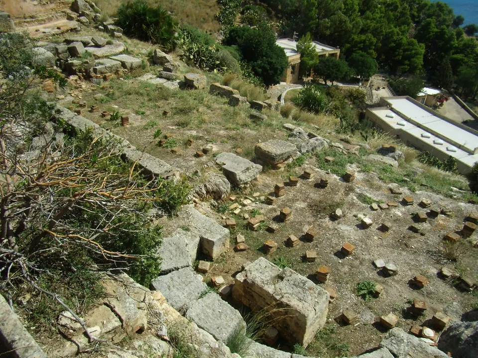 Looking down on an excavated floor set with rows of small square stones, framed by large cut blocks and low walls, with the trees and flat roofs of modern buildings beyond.