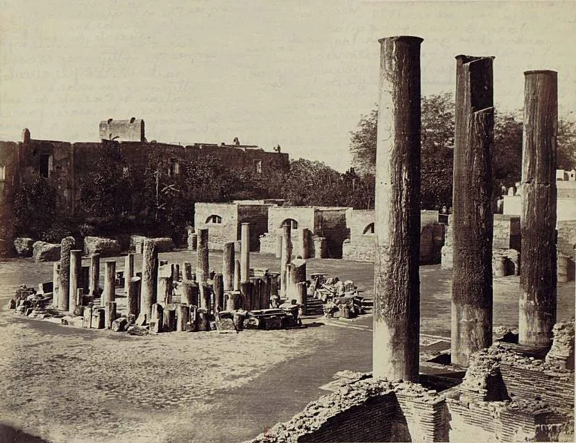 An old sepia photograph of three tall, weathered columns standing on the right, with rows of broken column stumps and low ruined buildings across dry ground behind them.