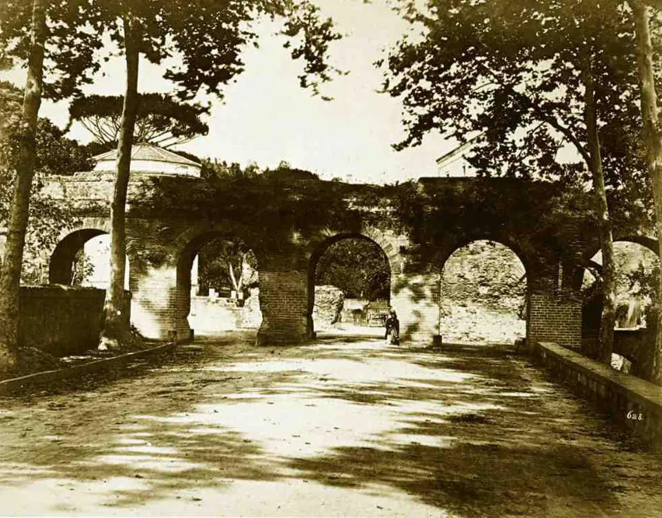 A sepia photograph of a ruined arcade of brick and stone arches at the end of a tree-lined road, with a small figure standing under one arch.
