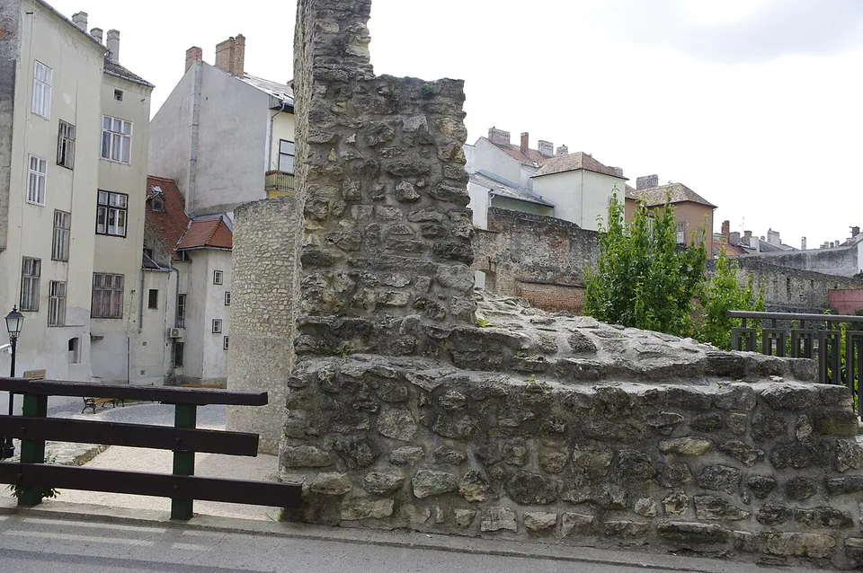 A rough stone wall, tall at one end and stepping down to a low wall top beside a railing, with old town houses behind.