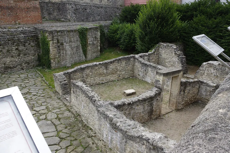 Low walls of rough stone forming small excavated rooms in a paved court below a taller old wall, with two information boards on stands.