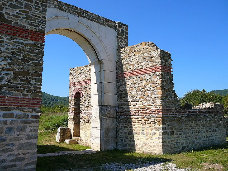 A re-erected round arch of white stone blocks set in a restored wall of rough stone banded with red brick, with a squat tower of the same masonry behind it and green wooded hills under a blue sky.