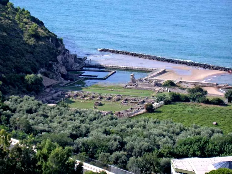 Seen from the hillside above, the low walls of rows of rooms laid out across green terraces run down to the shore, where rectangular stone-edged pools of seawater sit beside a beach and a breakwater at the foot of a wooded cliff, with trees in the foreground.