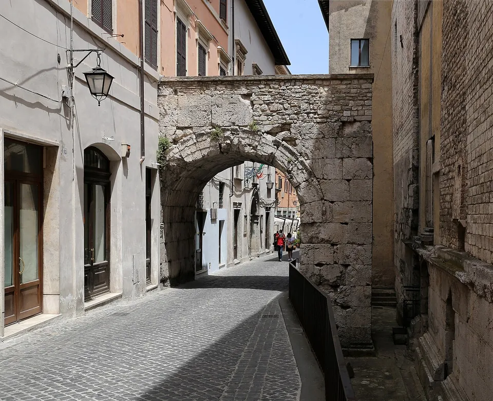 A single round arch of large rough limestone blocks spanning a narrow cobbled street between plastered houses, one pier merging into the house fronts and the other standing behind a railing, with people walking beneath.
