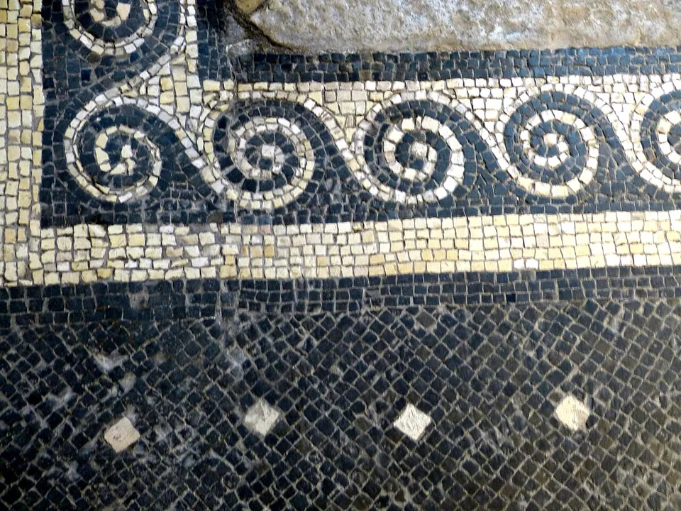 Close-up of a mosaic floor, a border of white running spirals on black between white bands, above a black field set with small white diamonds.
