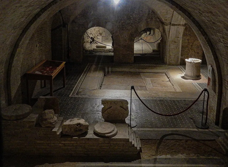 A dim underground hall under brick vaults, with a sunken square basin edged in stone in a dark mosaic floor, a fluted well-head beside it, a glass display case on the left, carved stone fragments in the foreground and more rooms through arches beyond, all behind rope barriers.