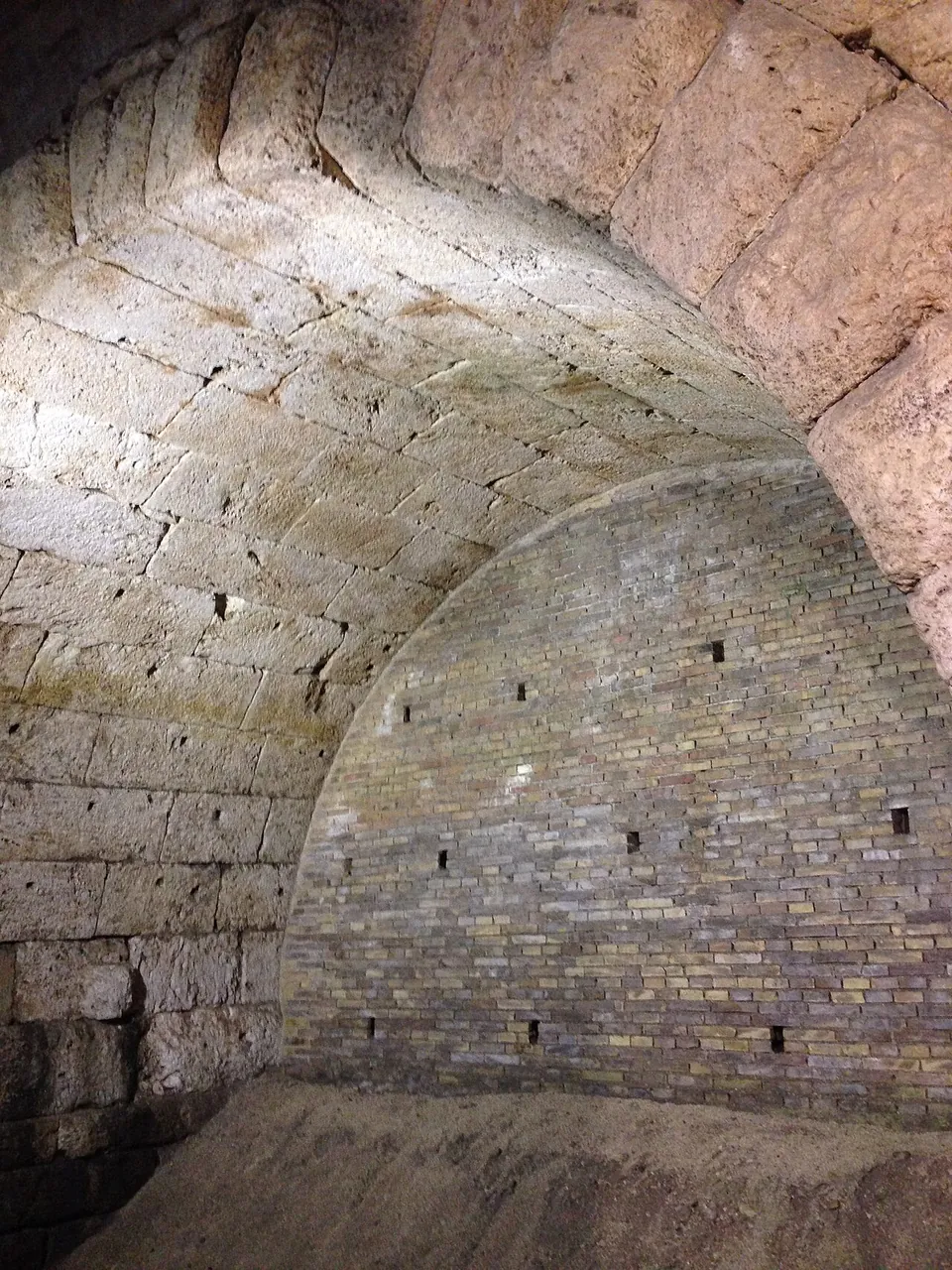 The underside of a vault of large squared stone blocks in artificial light, its far end filled with later brickwork.