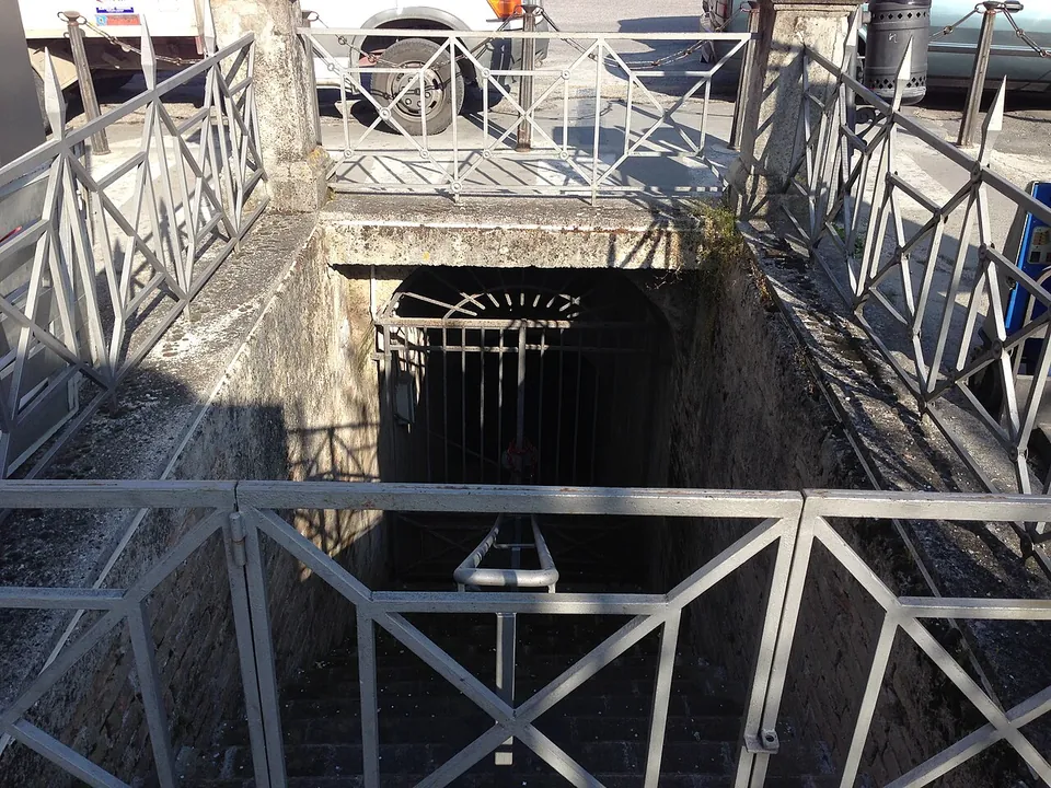 A rectangular opening in the pavement of a square, fenced with grey metal railings, with a stone stair leading down to an iron gate below street level and parked cars behind.