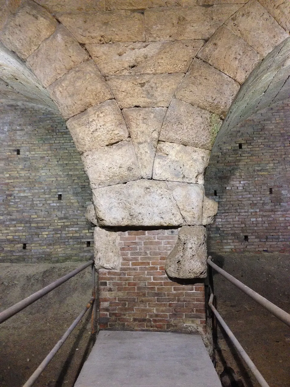 A pier of large, rough squared stone blocks rising into the springing of two vaults, its lower part filled with brickwork, seen in artificial light from a paved platform fenced by metal rails.