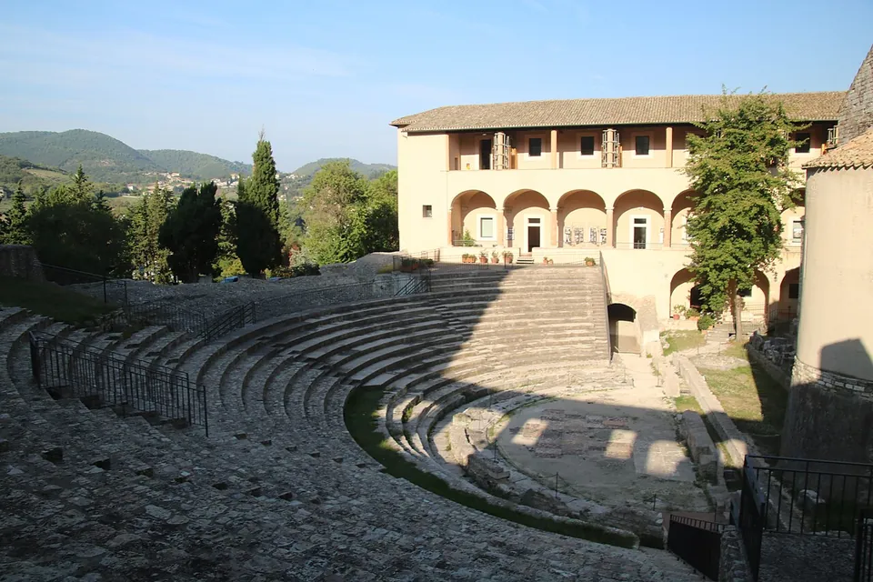 The stepped stone seating of a Roman theatre curving round a semicircular orchestra, with a long two-storey arcaded building and cypresses beyond it and green hills on the horizon.