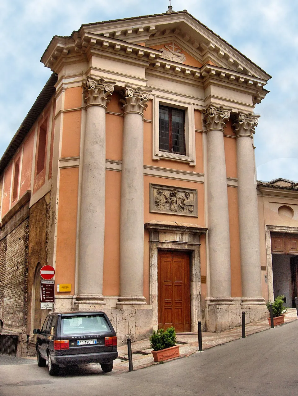 A church front with four tall columns carrying a triangular pediment, a carved relief panel over the door, peach-coloured plaster, and a car parked at the corner of a sloping street.