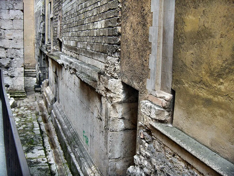 A narrow sunken strip below a street railing, running along the side of a church whose lower wall is built of large squared blocks with a moulded course, and plastered and brick walls above.