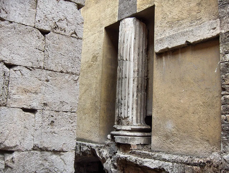 A fluted stone column standing in a tall recess of a plastered wall, its base on a moulded stone course, beside a wall of large squared limestone blocks.