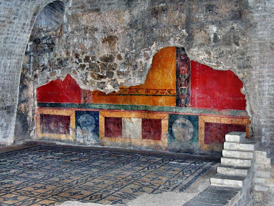 Wall painting at the foot of a rubble wall, with panels of deep red, ochre and green above a dado of coloured rectangles and roundels, over a mosaic floor, and a short flight of modern steps on the right.