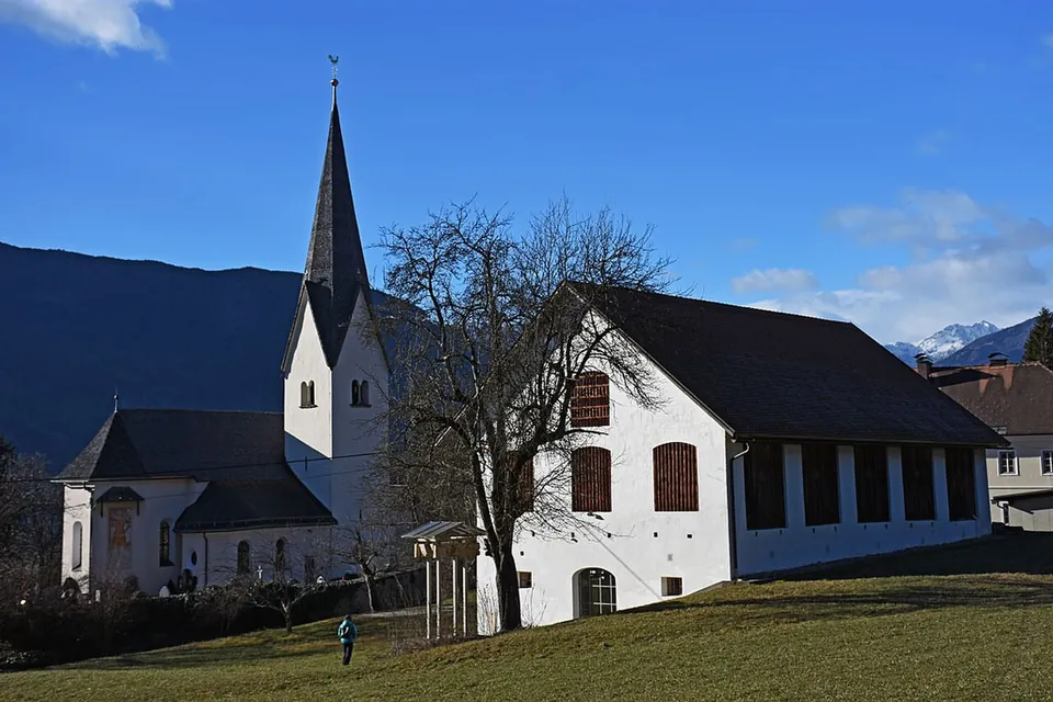 A white village church with a tall pointed spire next to a long white building with a dark roof and tall windows, on a grassy hillside under a blue sky.