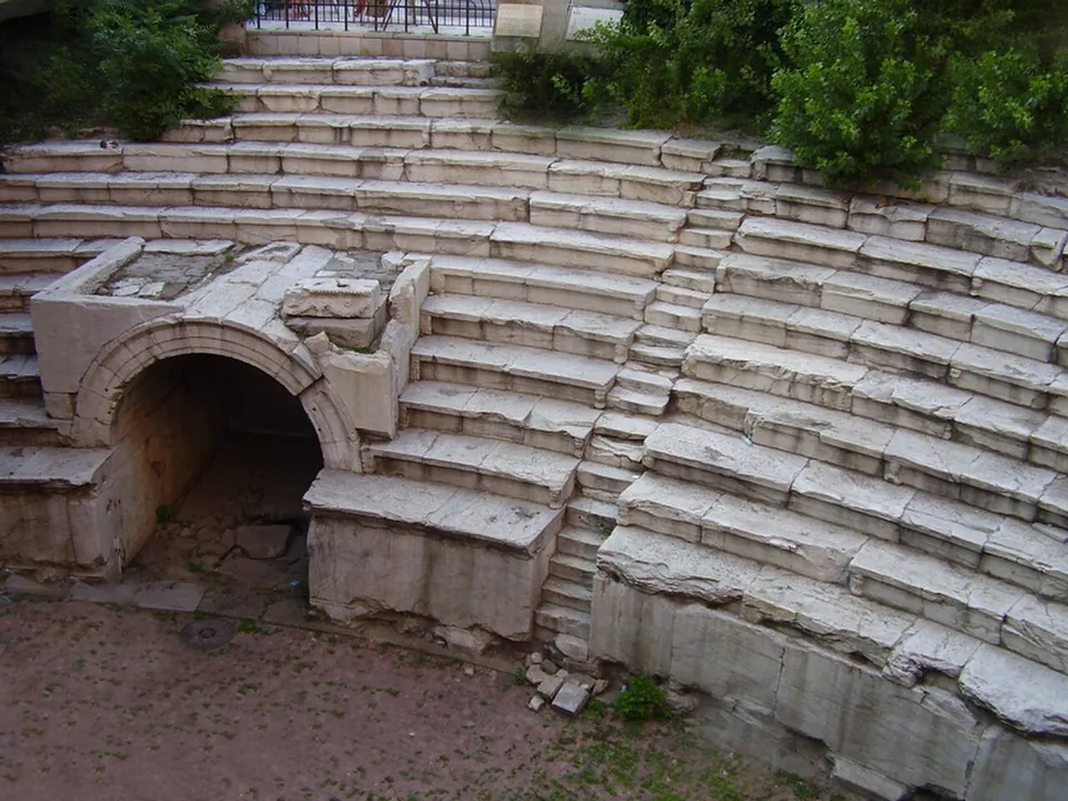 Tiers of worn white marble seats curving round a sunken floor, with a round-arched vaulted entrance set into the lower rows and shrubs at the top.