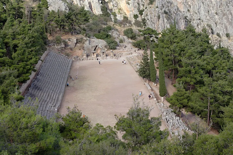 A long flat earthen running track seen from high above, with tiers of grey stone seats along one side, pines on the slopes around it and a cliff above.