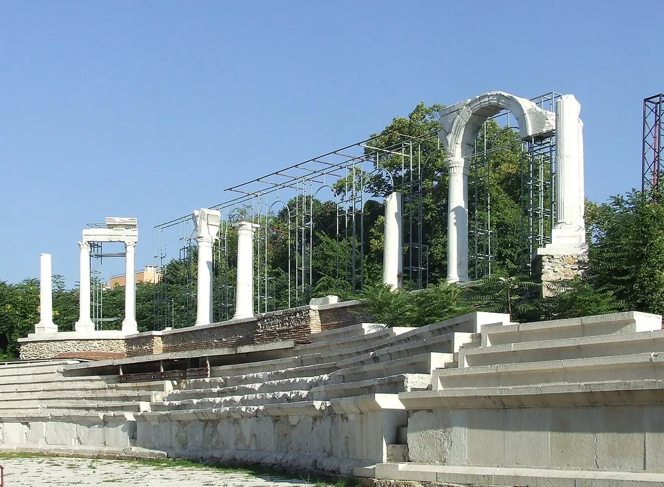Rows of pale stone seats rising in a shallow curve, with a line of re-erected white columns and a single tall arch along the top, partly held by modern metal scaffolding, under a clear blue sky.