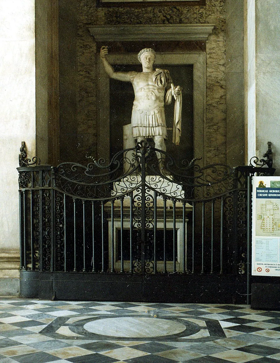 A marble statue of a man in Roman armour with his right arm raised, standing in a tall niche behind a wrought-iron railing, with an information panel at the right.