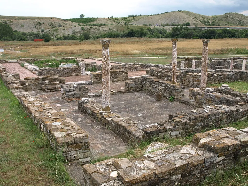 Low walls of stone courses around a paved court, with three tall columns still standing, dry grassland beyond and bare hills on the horizon.