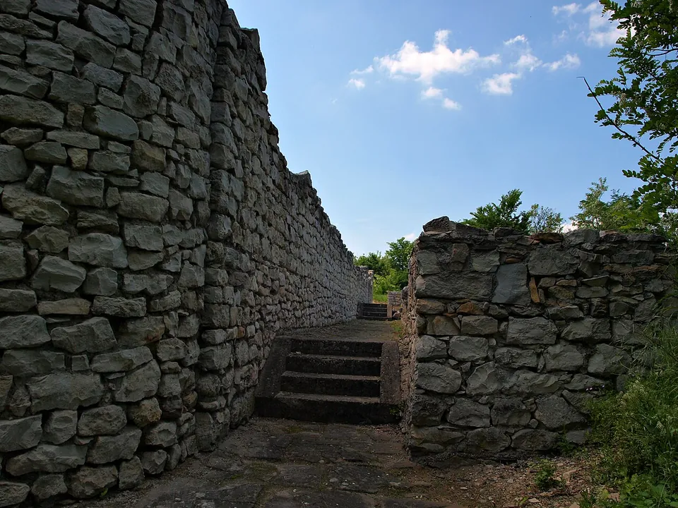 A narrow paved passage between a high wall of roughly squared stone and a lower wall, leading to a short flight of stone steps.