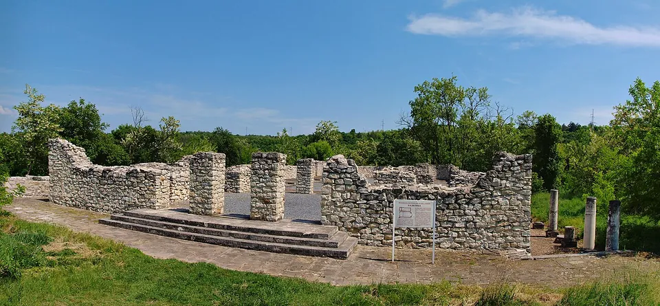 Restored walls of grey rubble stone enclose a paved rectangular space reached by a short flight of steps, with an information board in front, column shafts standing to the right and woods behind.