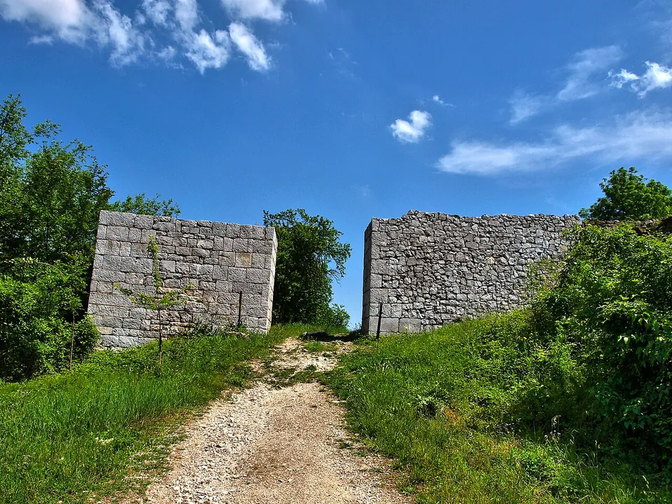 A gravel path climbs a grassy slope through a gap between two tall blocks of stone wall, with trees on either side and a blue sky.