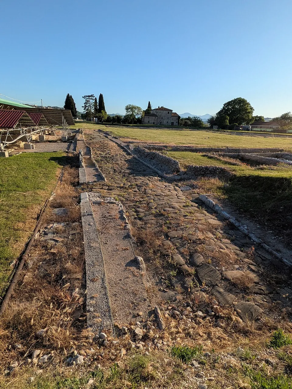A straight excavated road surface of stones and slabs, edged with grass, running towards a farmhouse and cypress trees, with the edge of a red roof at the left.