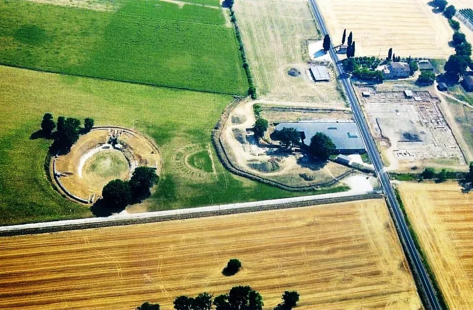 An aerial view of flat farmland in a valley. At the left an oval earthwork ringed with trees encloses a grassy arena; in the middle a long low roof stands among trees beside a farm track; a straight road runs along the right-hand side past an area of excavated ground.