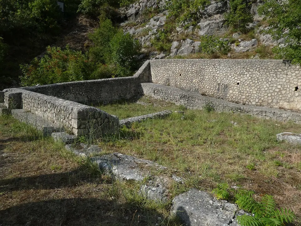 Terrace walls of small rounded stones set in mortar, stepping up a grassy slope in two levels, with scattered stone blocks in the grass and a rocky, wooded slope behind.