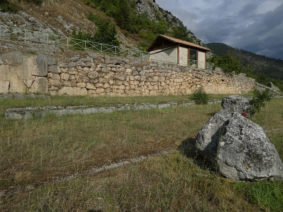 A large boulder lying on a grassy terrace below a long retaining wall of big stone blocks, with a small tiled-roof hut and a railing on the slope above.