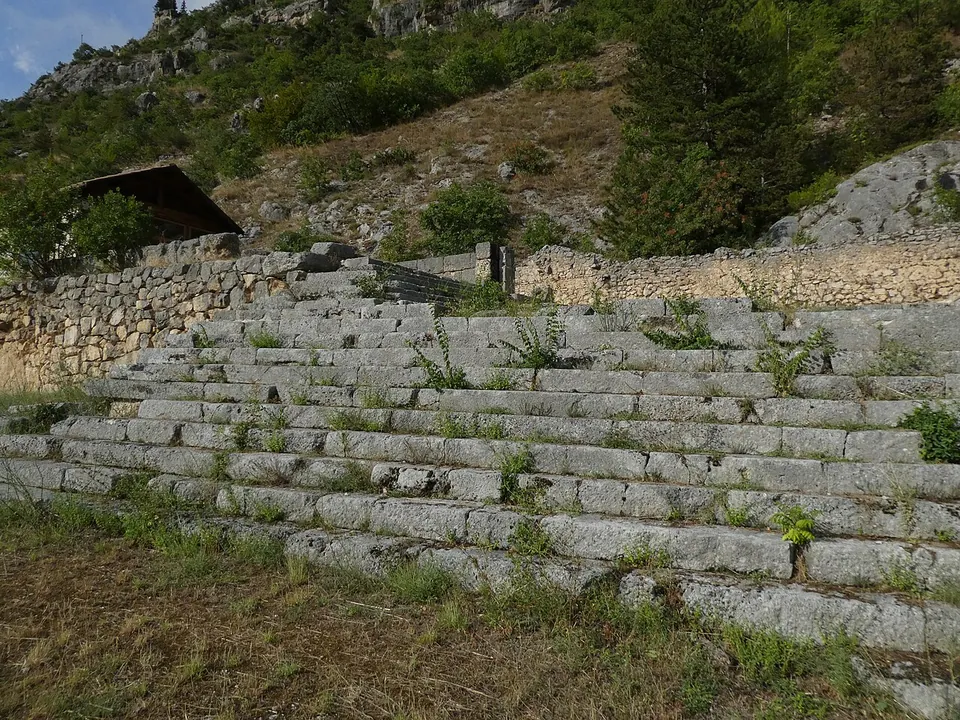 A broad flight of grey stone steps, many treads wide, climbing a grassy slope to a wall of mortared stone, with a small tiled-roof hut and a wooded, rocky mountainside above.
