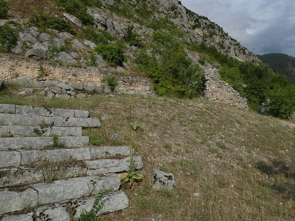 Stone steps at the edge of a dry grassy slope, with low ruined walls beyond and bare limestone slopes rising under a cloudy sky.