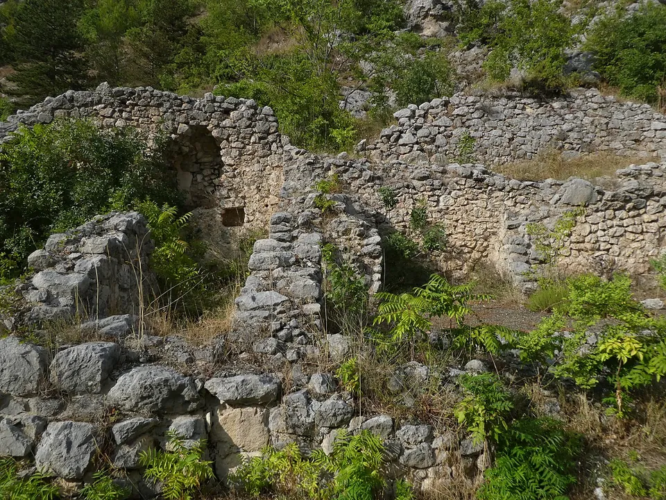 Ruined walls of rough stone rubble, overgrown with ferns and shrubs, with a small arched opening in the wall at the back.