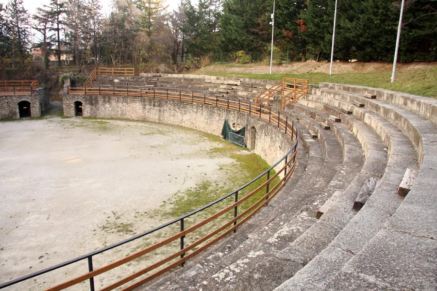 An oval arena of bare, pale earth ringed by a low stone wall with small arched openings in it, and on one side rows of stone seating behind a metal railing, with a timber platform and tall conifers beyond.