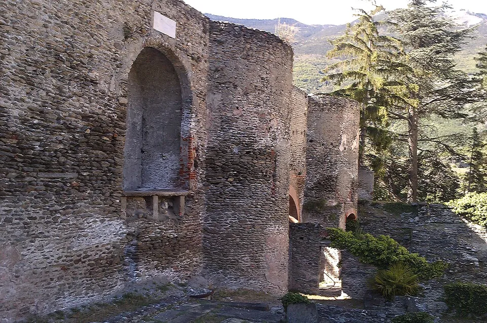 A tall wall of rough rubble stone with a round-headed niche in it, a rounded tower beside it and further stretches of wall beyond, with conifers and mountains behind.