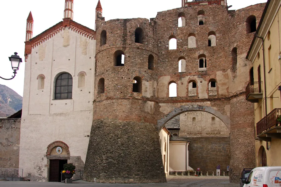 A tall gate of rubble stone and brick, two round towers with a sloping stone base and rows of arched window openings above, joined over a wide arched passage, with the white gabled facade of the cathedral standing against it on the left.