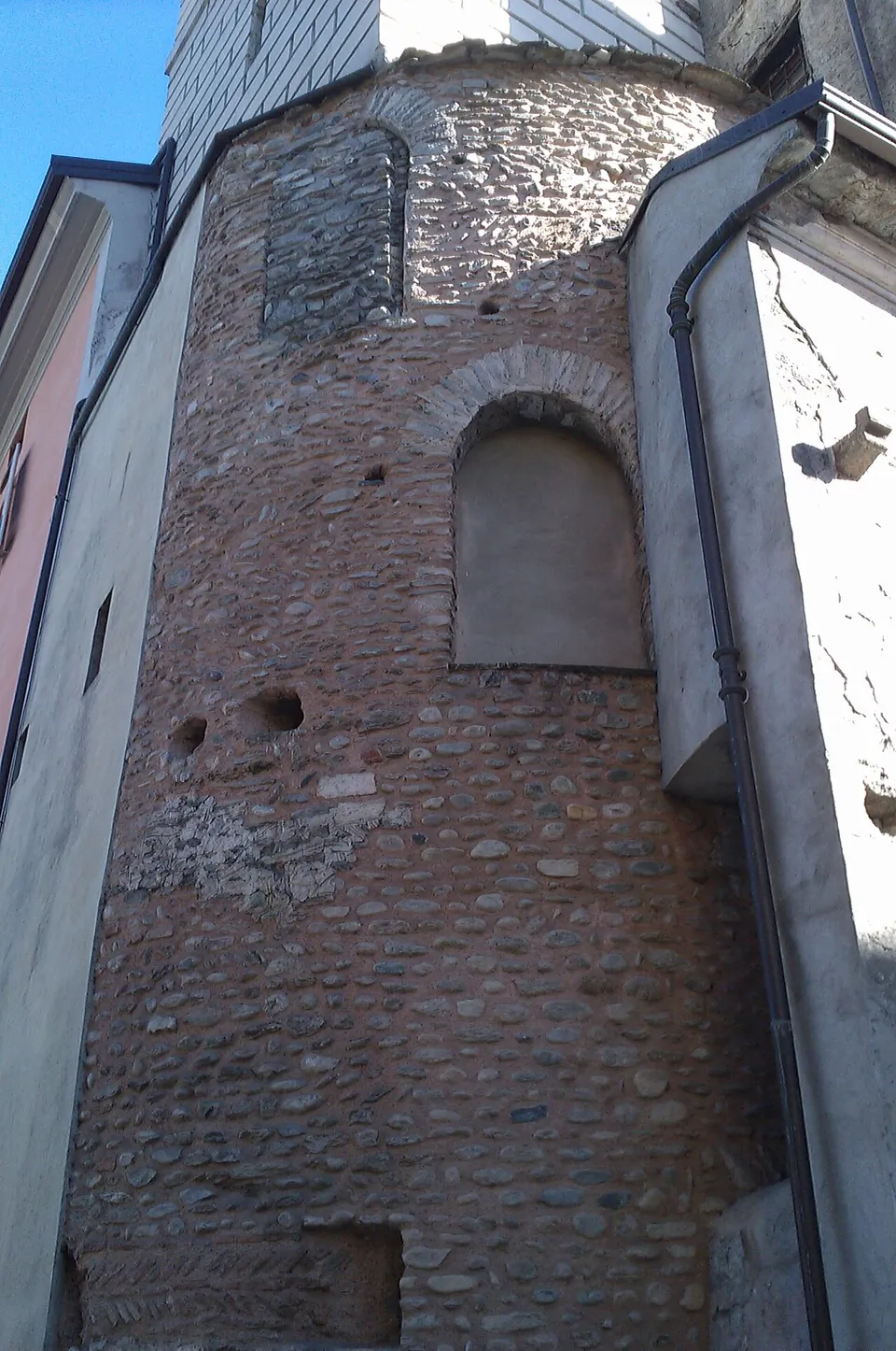 The upper part of a round tower of brick and rubble stone with blocked arched windows, built in among plastered houses, seen from below against a blue sky.