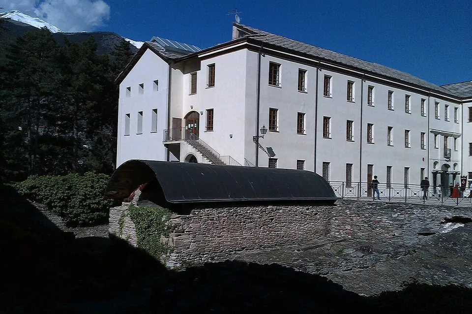 A long white three-storey building with an outside stair, and in the shade in front of it low stone walls under a curved dark roof.