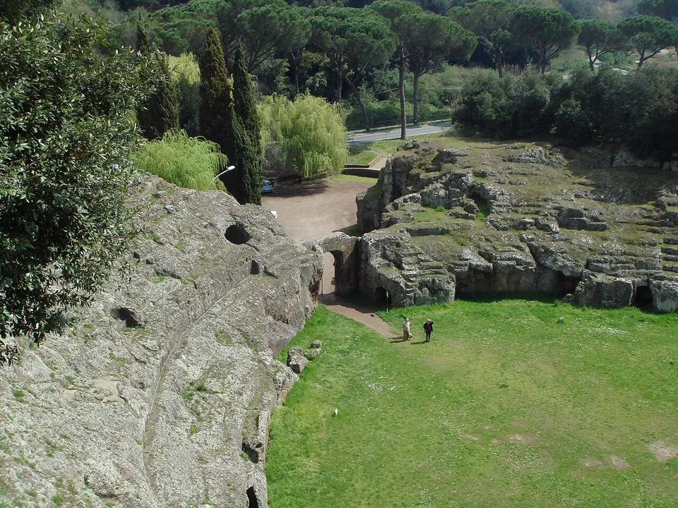 Two people walking across the grass arena towards an entrance passage cut through the rock, with the eroded seating on either side and a gravel area and road beyond the entrance.