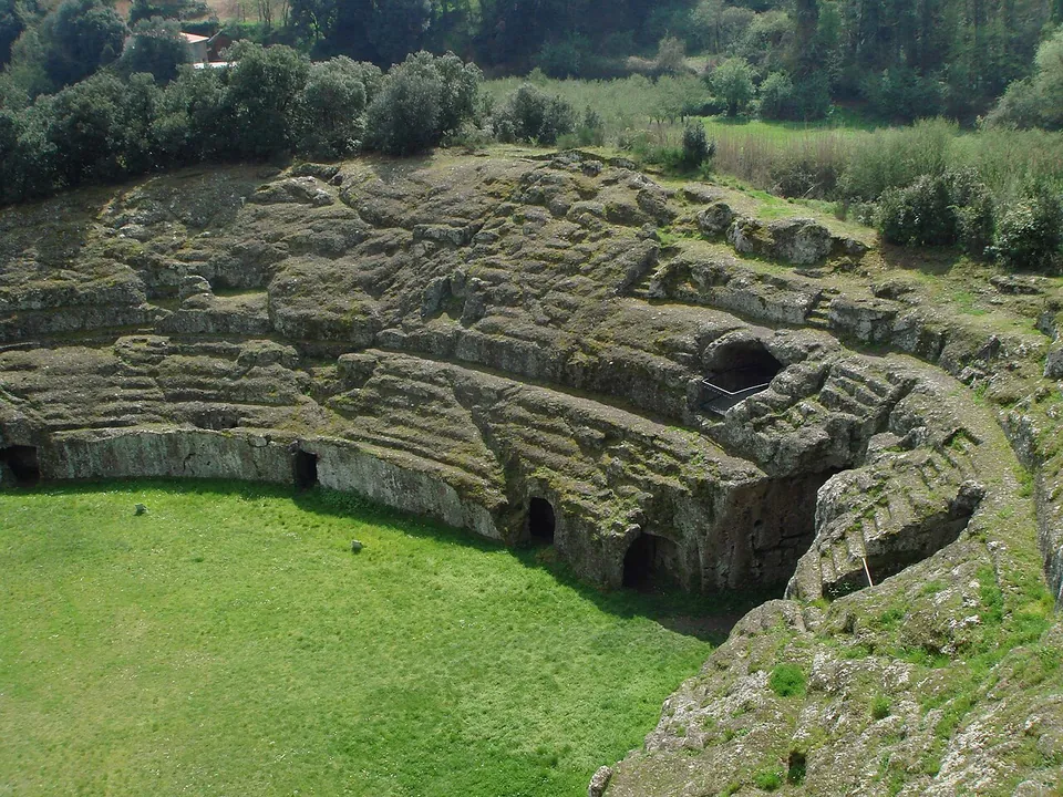 Looking down on one curve of the amphitheatre, where tiers of seating carved out of the grey-green tuff rise above a mown grass arena, with dark openings cut into the rock wall at arena level and trees on the ridge behind.