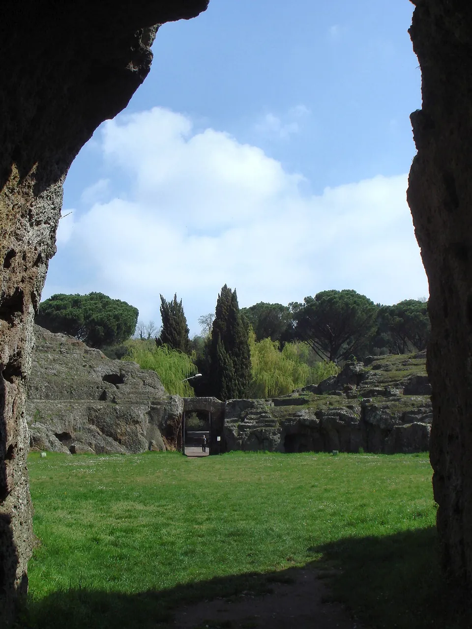 The arena framed by the dark rock walls of a passage, with the far side of the seating, cypresses and umbrella pines beyond under a blue sky.