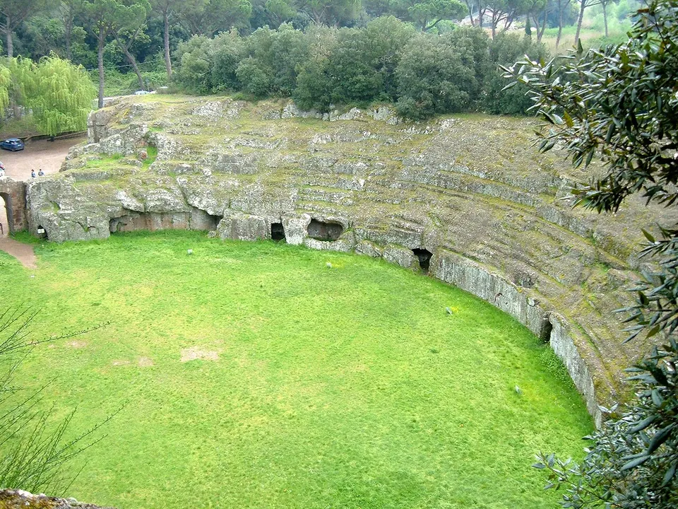 The whole oval arena from above, a lawn ringed by a rock wall pierced with low doorways and by worn tiers of seating, with woodland all round.